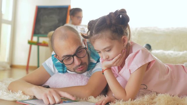 A man with a daughter pre-teen lie on the floor together and read a book, and in the background the eldest daughter on the couch leads video blog.