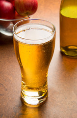 A glass of cold apple cider on a brown table with a glass bottle and ripe red apples. Selected focus.