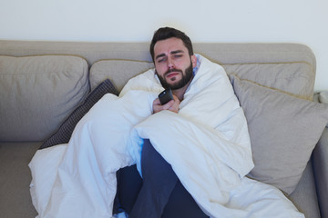 Sick young man with remote control sitting on couch in front of tv set