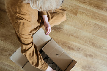 A young woman who is a customer prepares a package for shipping using tape and scissors. Girl consumer holding cardboard box on the floor.