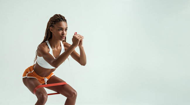 Fitness trainer. Full length of young and slim african woman in sports clothing exercising with a resistance band in studio against grey background