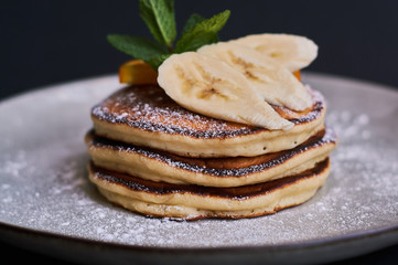 Homemade breakfast of pancakes decorated with orange, banana, mint and sugar powder on black background in grey plate.