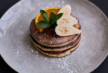Homemade breakfast of pancakes decorated with orange, banana, mint and sugar powder on black background in grey plate.