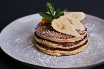 Homemade breakfast of pancakes decorated with orange, banana, mint and sugar powder on black background in grey plate.