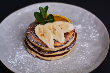 Homemade breakfast of pancakes decorated with orange, banana, mint and sugar powder on black background in grey plate.