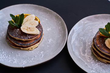 Homemade breakfast of pancakes decorated with orange, banana, mint and sugar powder on black background in grey plate.
