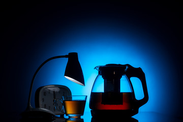 glass teapot and cup with hot tea and table lamp decorative with light garland on dark blue background, close view  