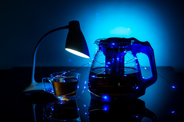 glass teapot and cup with hot tea with table lamp on dark blue background, close view  