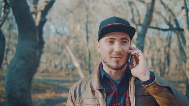 Young man in the autumn park. The guy in the leather jacket and baseball cap is talking on the smartphone. The guy on the background of trees without foliage.