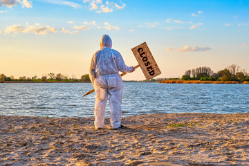 Man wearing protective suit stands with warning sign in hands with inscription closed along the sand beach of the river. Contaminated water, quarantine, virus outbreak, crime scene concept