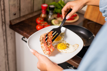 Hands of young female putting fried bacon and eggs on plate in the kitchen