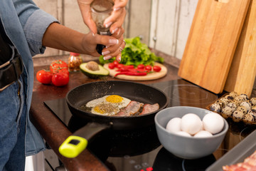 Hands of young woman sprinkling spices on fried bacon and eggs during cooking