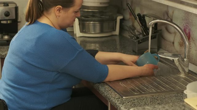 Disabled woman in a wheelchair washes dishes in the kitchen in the wash basin