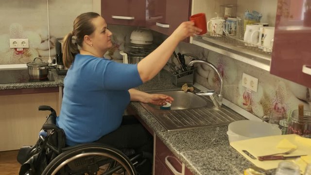 Disabled woman in a wheelchair washes dishes in the kitchen in the wash basin