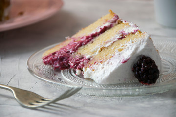 Close up of a sponge cake portion filled with whipped cream and berries sauce with a blackberry as a decoration on a crystal plate and a fork. Naked cake concept.