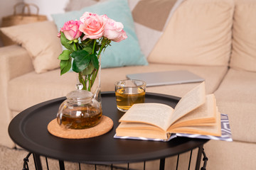 Small table with rose bunch in glass of water, green herbal tea and open book