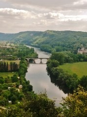 Fotografía de una típica estampa de la idílica región de la Dordogna francesa con un pequeño castillo rodeado de diferentes tonos de color verde del bosque y un puente de arcos cruzando un río