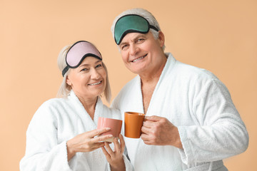 Mature couple in bathrobes and with sleep masks drinking tea on color background