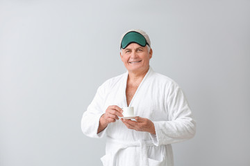 Happy mature man in bathrobe, with sleep mask and cup of coffee on grey background