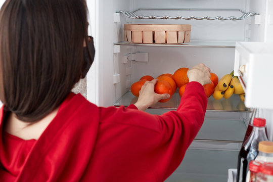 Back view of woman wears red costume, stands in front of opened refrigerator, chooses fruits for making fresh salad. People, eating and nurtrition concept