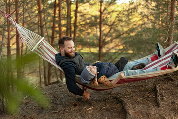 Young family hipster playing with son on a hammock in park on camping trip, family vacation