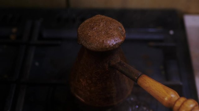 Close-up of boiling coffee escapes from the narrow neck of small copper pot with wooden handle and extinguishes the blue fire on the gas stove