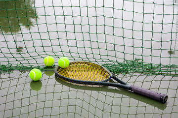Tennis balls and racket on wet tennis court after raining. Racket and tennis balls laying beside a net on court covered with rain..