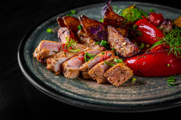 fried meat with potatoes, pepper, tomatoes, herbs and spices in plate on black wooden table background