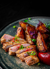 fried meat with potatoes, pepper, tomatoes, herbs and spices in plate on black wooden table background