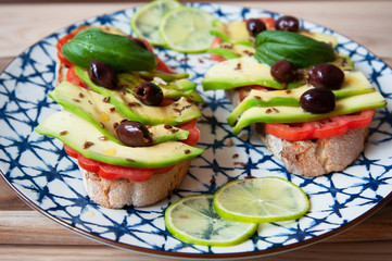 Avocado sandwich toast on ceramic vintage dish. Fresh vegetarian ingredients used on brunch meal. Bruschetta, Avocado, Tomatoes, Olives, Flaxseed, Basil, Lime. Healthy lifestyle. Raw food concept.