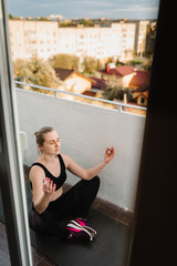 Woman sitting on the mat on balcony in yoga pose. Young girl practicing yoga at home on sunny day breathing fresh air. Calmness concept. Covid-19 Coronavirus Stay Home. Athlete trains during pandemic.