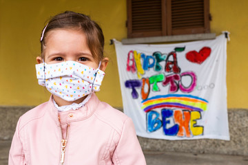 Little girl with homemade face protective mask. Blurred background with the famed rainbow drawing...