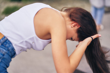 A girl in a white top and jeans, bending over, collects her hair in a ponytail.