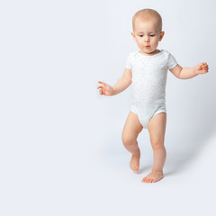 The first steps of the baby. The little girl walks unsteadily, splay-footed, spread her hands to the sides for balance. A child on a white background in a white bodysuit.