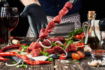 A butcher holds a skewer of fresh meat over four meat skewers that lie on a stone cutting board on an old village table. In the background, various condiments and spices, a glass and a bottle of wine.