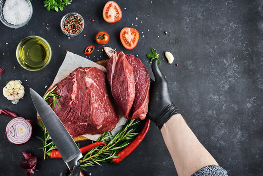 The butcher shows a section of fresh meat tenderloin, which lies on parchment on a dark background. On the table-seasonings, spices, vegetables and herbs. Top view, free space for text.