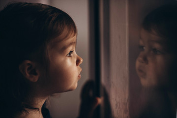 Toddler Caucasian cue girl in looking in the window in self-isolation during covid virus feeling...