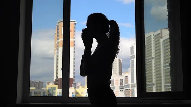 Epidemic Coronavirus Covid-19. Silhouette young woman at the window. Girl puts on a facial mask before going out. Life in quarantine self-isolation. Pandemic. Modern apartment buildings on background.