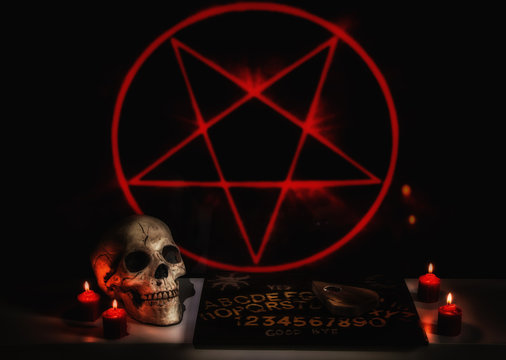 A satanic seance ritual scene with a skull, ouija board, planchette and red burning candles.
