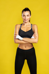 Fototapeta na wymiar Ready for workout. Young beautiful woman in sportswear keeping arms crossed and looking at camera while standing against yellow background