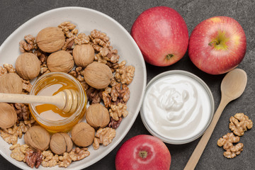 Walnut kernels and whole walnuts, glass jar with honey in white ceramic bowl, yogurt and red apples on table