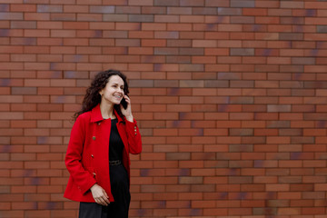 Smiling young woman in red, hair in wind, talking on the smartphone while walking outside, near brick wall. Cheerful woman using mobile. Space for text.
