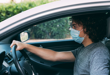 Young curly man driving a car in a medical mask and gray T-shirt. Man in protective mask is sitting at the wheel of a car. Protection from coronavirus epidemic pandemic, covid-19. New reality