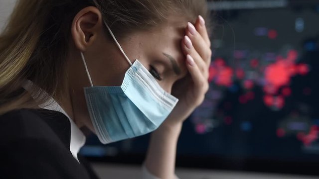 Side view close-up portrait of a young woman in disposable medical facial mask holding her head and looking at the epidemic map of the coronavirus Covid 19 pandemic on a computer