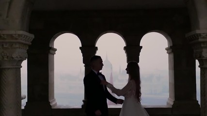 silhouette of young couple on window background of arches of old building, architecture, on city landscape background. the bride and groom look at each other, stand, embrace holding hands. Slow motion