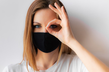 Beautiful close-up portrait of a young woman in a black medical mask and white T-shirt on white background, makes okay sign with hand. Quarantine, stop coronavirus pandemic, covid-19. Stay home.