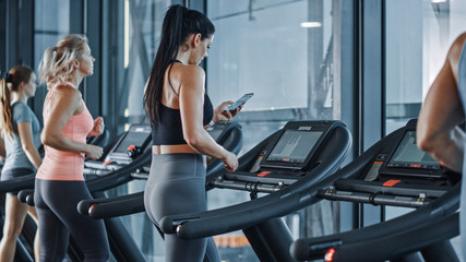 Fototapeta na wymiar Athletic Sports Woman in Gym Puts on Wireless Headphones, Turns on Sport Music Playlist with Smartphone and Starts Running on Treadmill. In Background Fit Athletes Training.