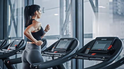 Fototapeta na wymiar Beautiful Athletic Sports Woman Wearing Wireless Headphones, Listens to Podcast or Sport Music Playlist while Running on a Treadmill. Energetic Female Athlete Training in the Gym Alone. Side Back View