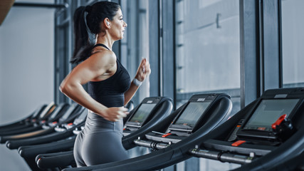 Fototapeta na wymiar Beautiful Athletic Sports Woman Wearing Wireless Headphones Listens to Podcast or Sport Music Playlist while Running on Treadmill. Energetic Female Athlete Training in Gym Alone.