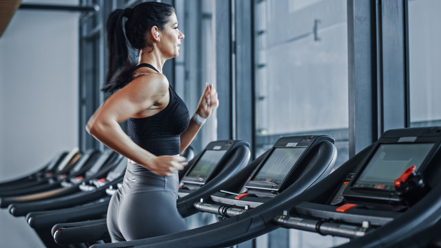 Beautiful Athletic Sports Woman Wearing Wireless Headphones Listens to Podcast or Sport Music Playlist while Running on Treadmill. Energetic Female Athlete Training in Gym Alone.